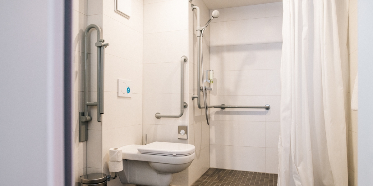 Barrierearmes Badezimmer des Doppelzimmers im Kongresshotel Potsdam. Helle Wandfliesen und dunkle Bodenfliesen. In der Mitte ist die Toilette mit seitlichen Halterungen und rechts eine offene Dusche ebenfalls mit Halterungen und einem weißen Duschvorhang.